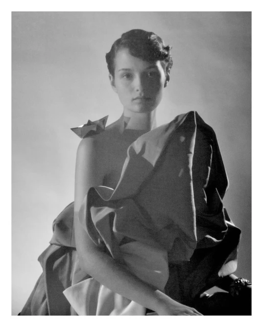 Monochrome portrait of a young woman with draped fabric and a sculptural paper detail, seated against a neutral backdrop in a studio setting.