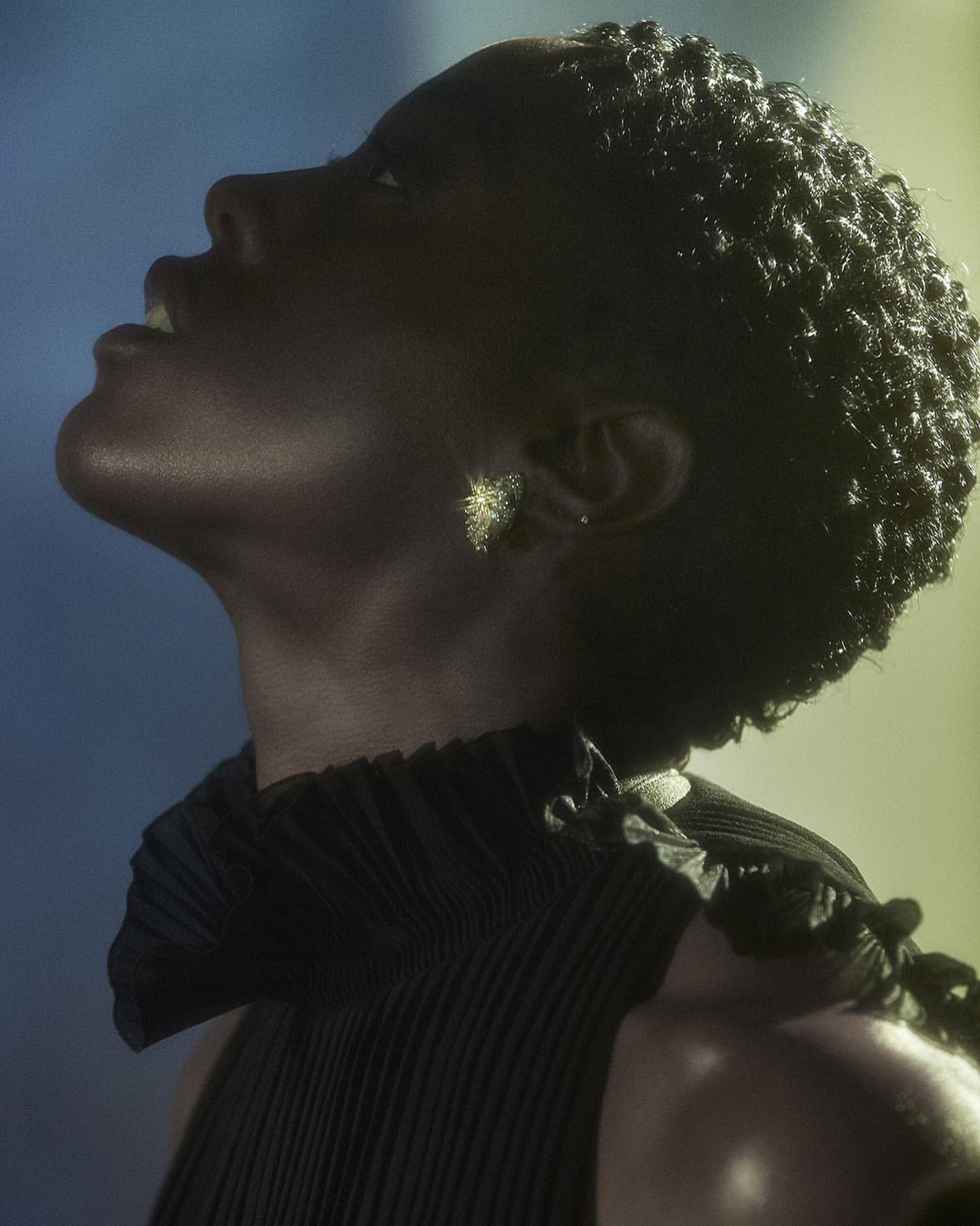 Portrait of Black woman with elegant updo and black pleated garment against a gradient blue and yellow backdrop in a studio setting.