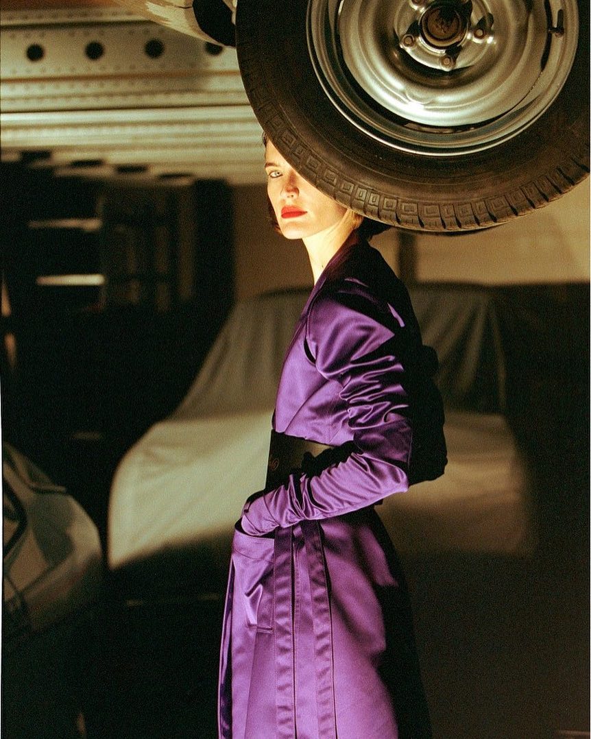 Elegant woman in purple dress poses beneath a car wheel in a stylized, high-fashion automotive-themed photoshoot with dramatic lighting.