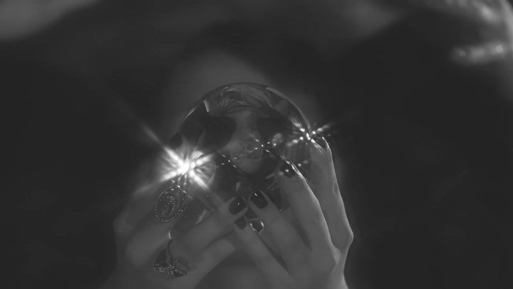 Close-up of a woman's hands holding a crystal ball, with rings and dark nail polish visible against a dark background in monochrome.