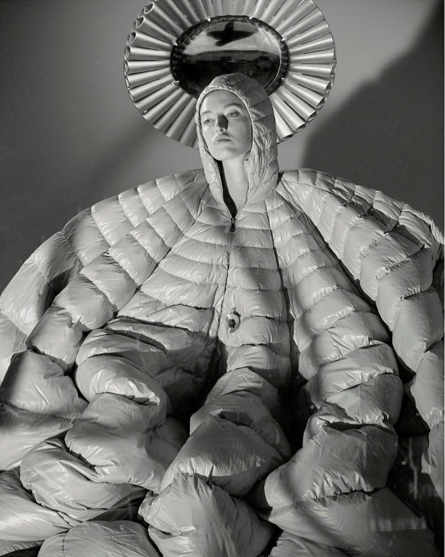 Monochrome fashion shot of a person posing in a large, quilted coat with hood, against a neutral backdrop with round mirror.