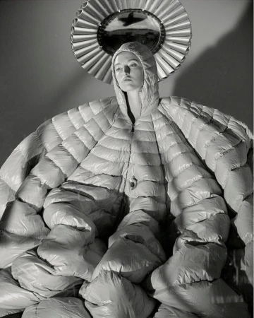 Monochrome fashion shot of a person posing in a large, quilted coat with hood, against a neutral backdrop with round mirror.