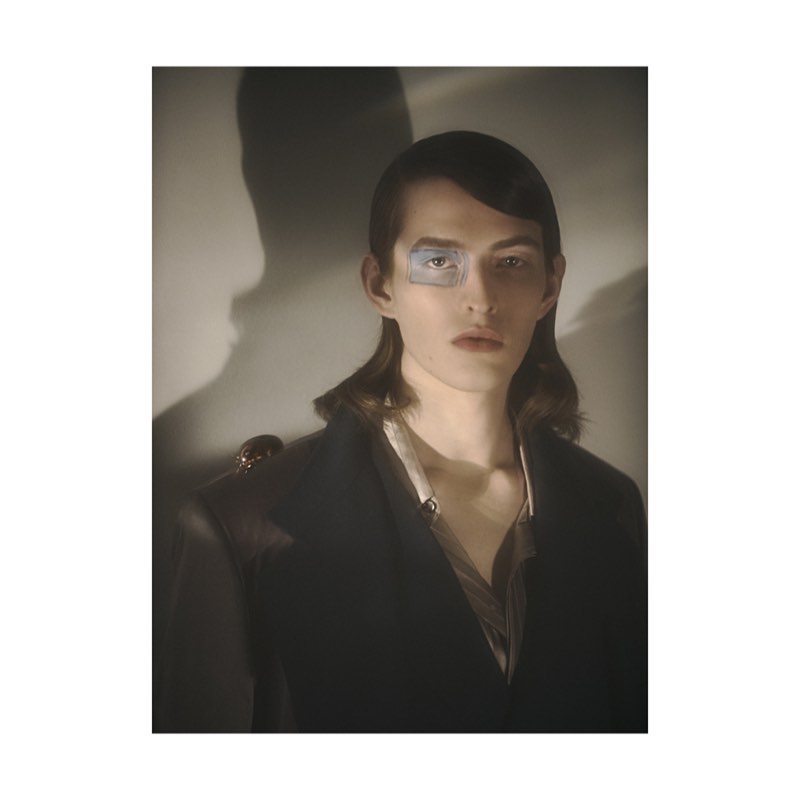Portrait of a man with dark shoulder-length hair wearing a dark jacket against a light wall with a prominent shadow.