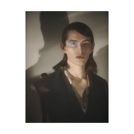 Portrait of a man with dark shoulder-length hair wearing a dark jacket against a light wall with a prominent shadow.