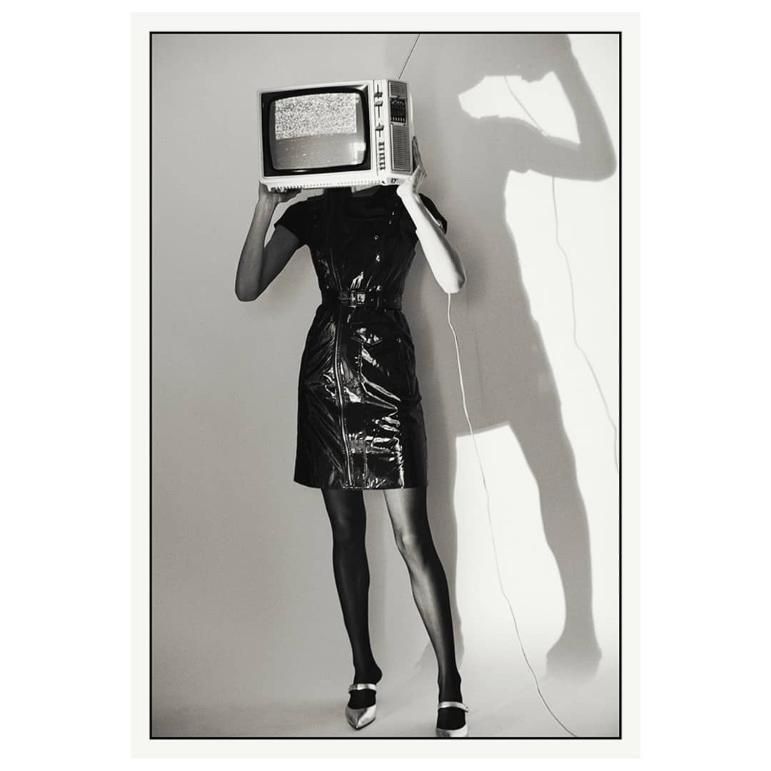 Monochrome image of a woman wearing a black patent leather dress and silver heels holding an old television over her head.