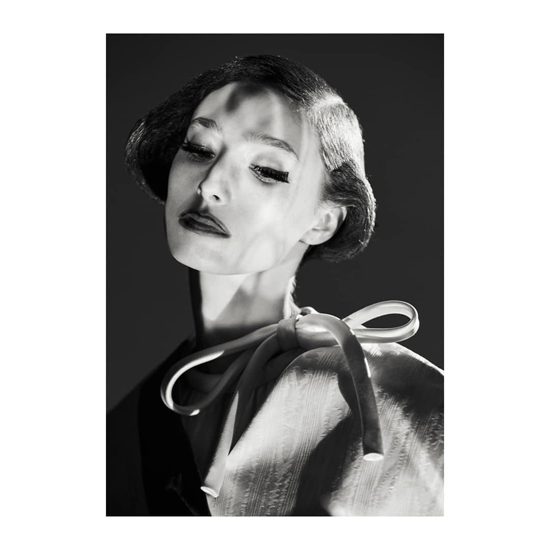 Monochrome portrait of a stylish adult woman with short, dark, wavy hair, wearing an off-white blouse with a ribbon bow. Dramatic lighting.