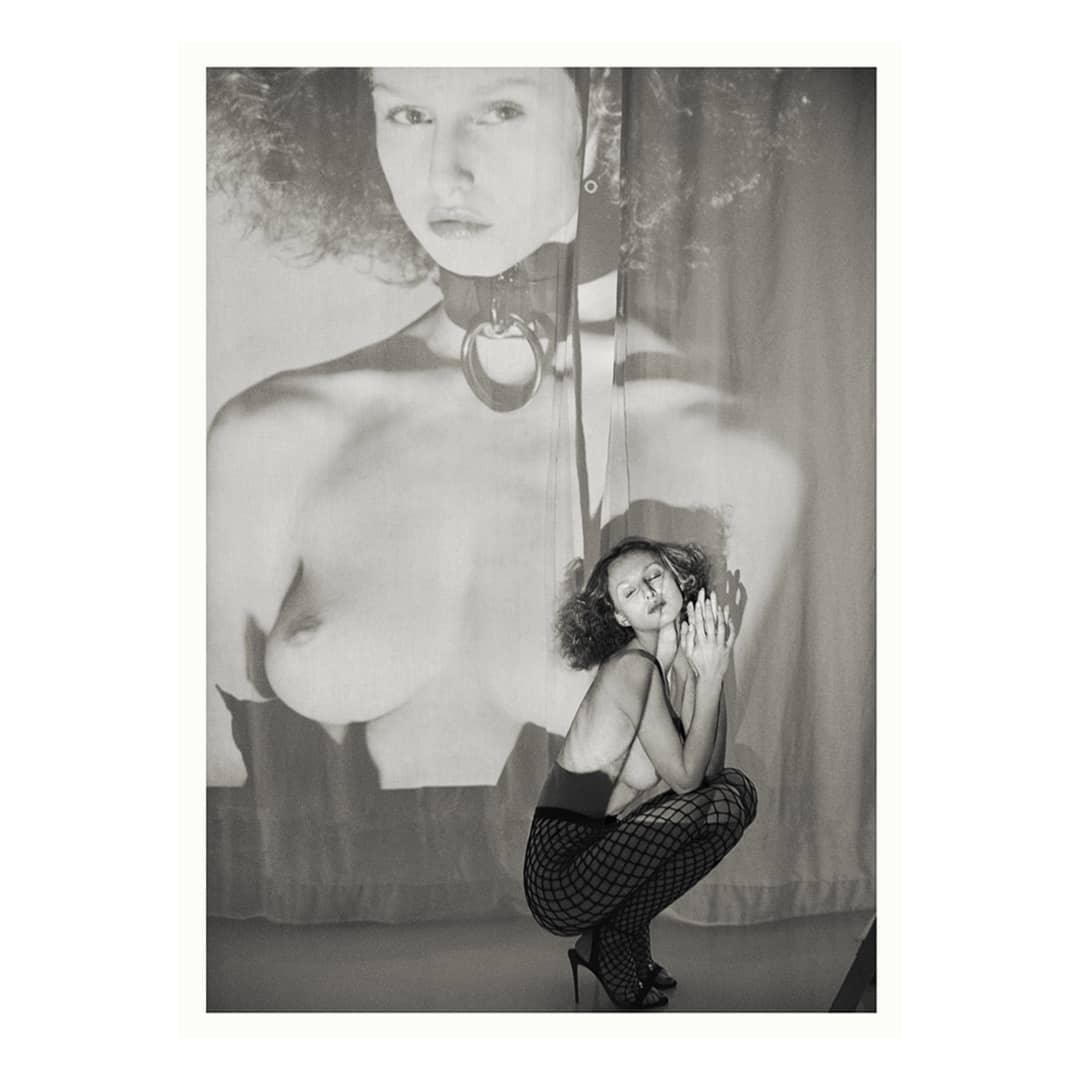 Monochrome image of a woman crouching in fishnet stockings and high heels, with a projected image behind her creating a layered effect.