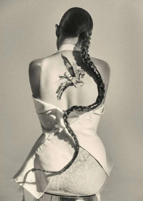 Back view of a woman with a long braid, wearing a white top, and a Pegasus tattoo on her back in a studio setting.