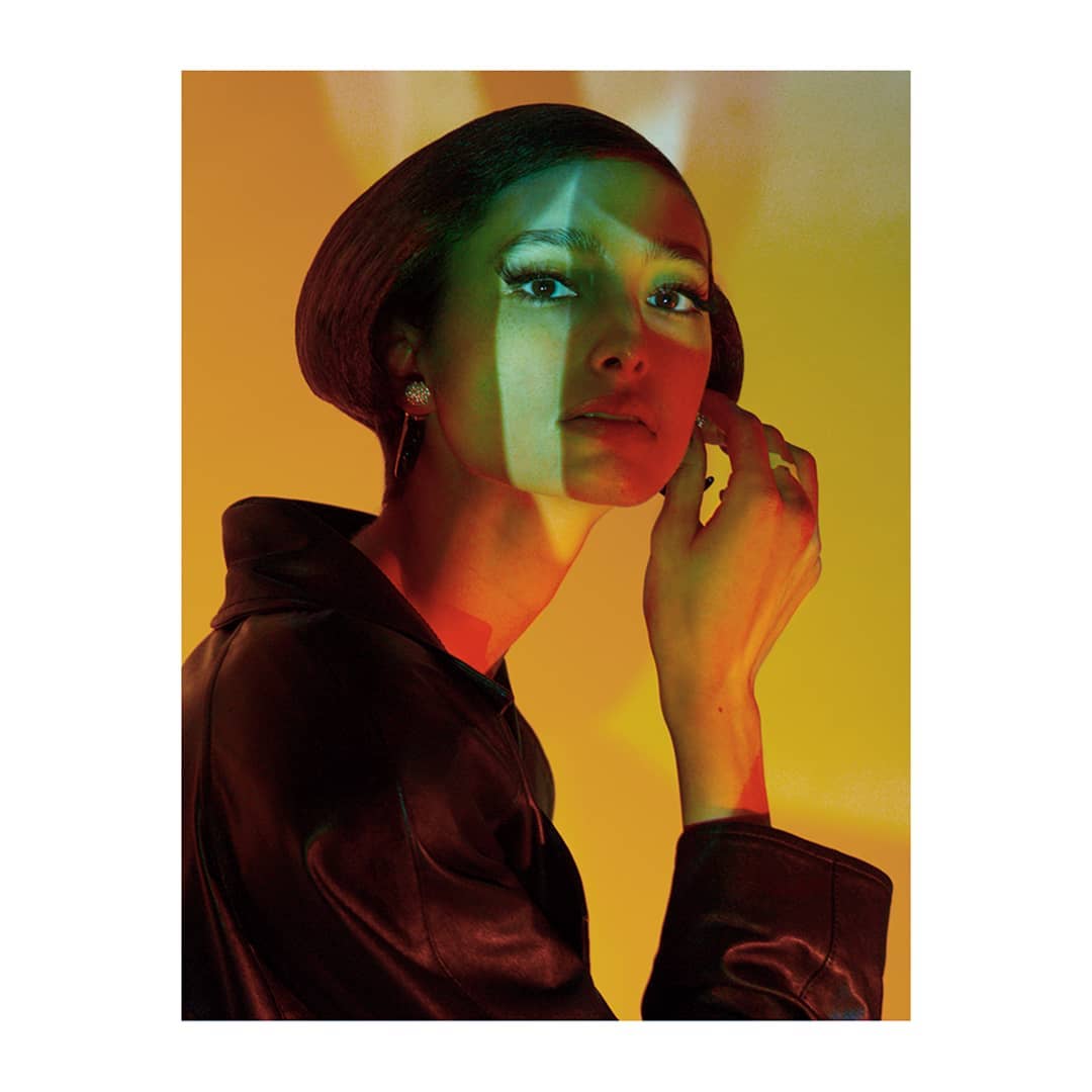 Elegant woman in a leather jacket stands in a studio with dramatic lighting creating green and red shadows on her face and yellow background.