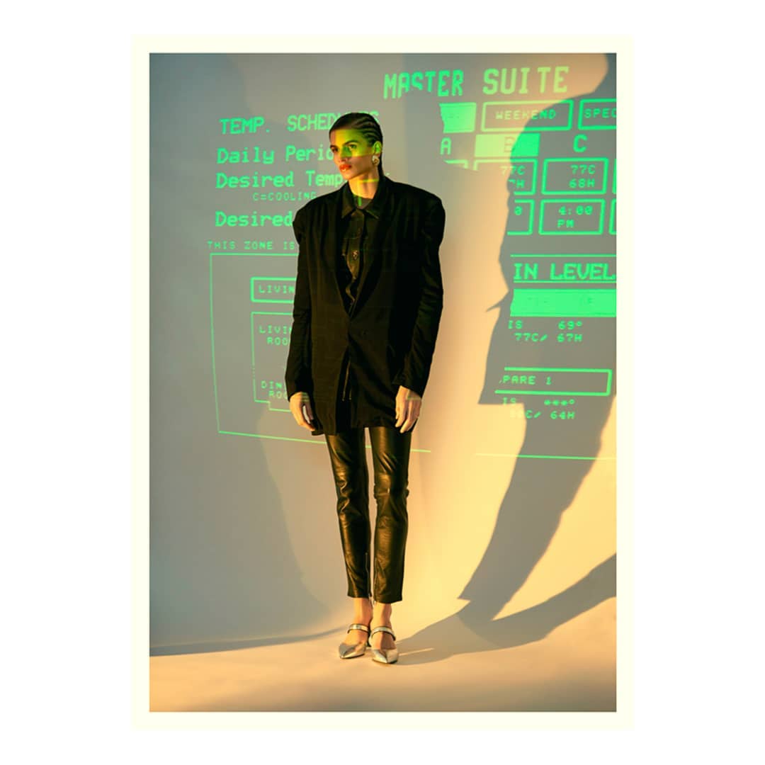 A woman poses in a black blazer and leather pants against a green-lit computer display in a studio setting, casting a long shadow.