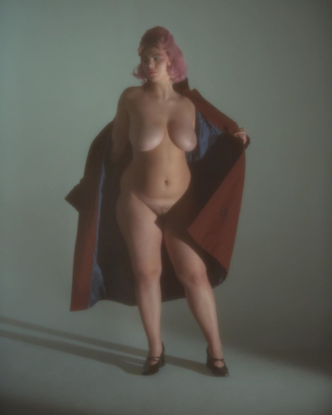 Topless woman with pink hair and a brown coat standing in a light-grey photo studio with a blurred background and soft lighting.