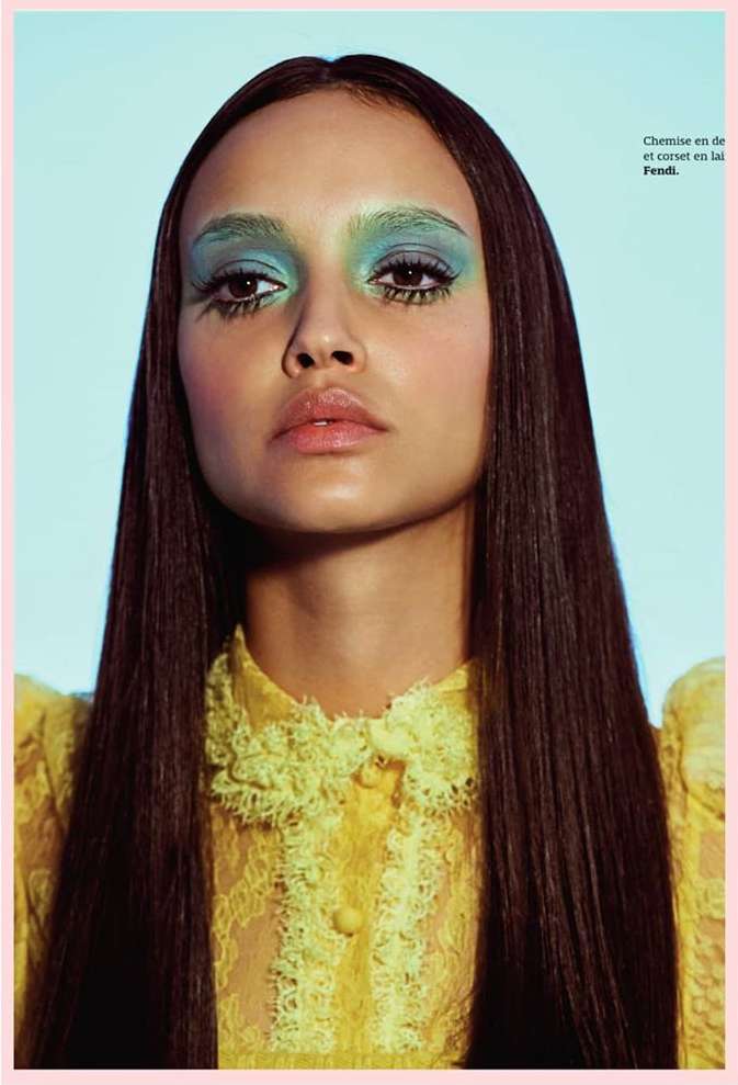 Young woman with dramatic turquoise eye makeup and sleek brunette hair poses in front of a light blue background wearing yellow lace top.