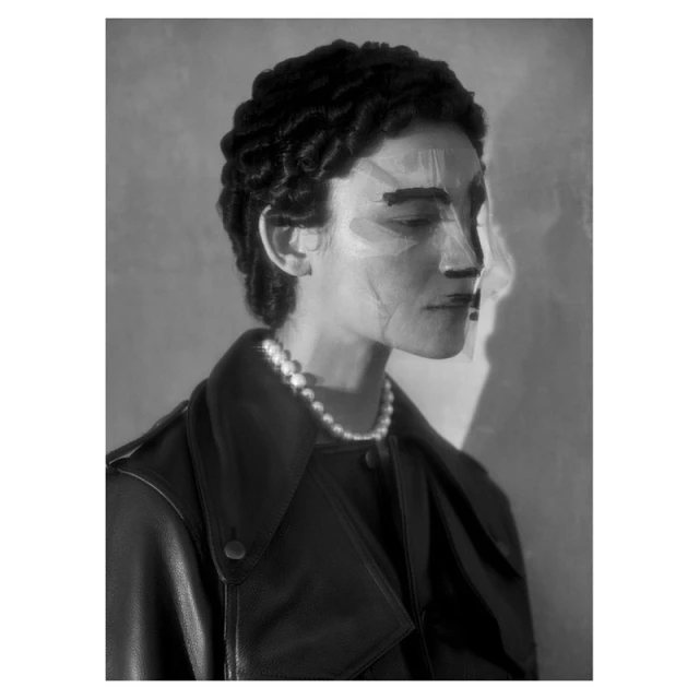 Monochrome portrait of a woman with stylized makeup wearing a leather jacket and pearl necklace against a diffused, neutral background.