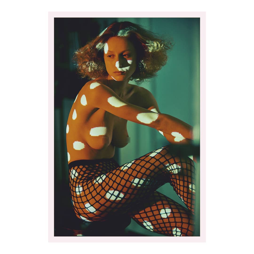 A topless woman in fishnet stockings poses in a studio, with a projection of white dots covering her body, creating a patterned effect.
