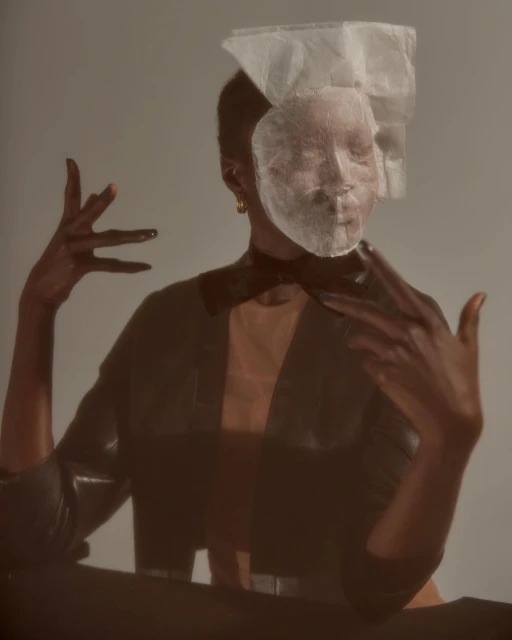 Artistic portrait of a woman with short dark hair wearing a jacket, obscured by a translucent face covering and expressive hand gestures.