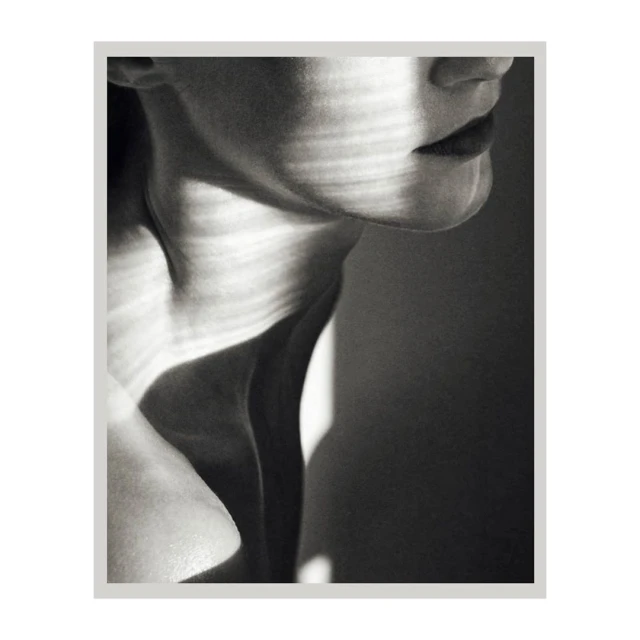 Monochromatic studio portrait of a model’s shoulder and neck with shadows creating graphic patterns on her skin in a high-key setting.