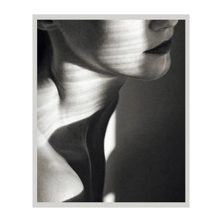 Monochromatic studio portrait of a model’s shoulder and neck with shadows creating graphic patterns on her skin in a high-key setting.