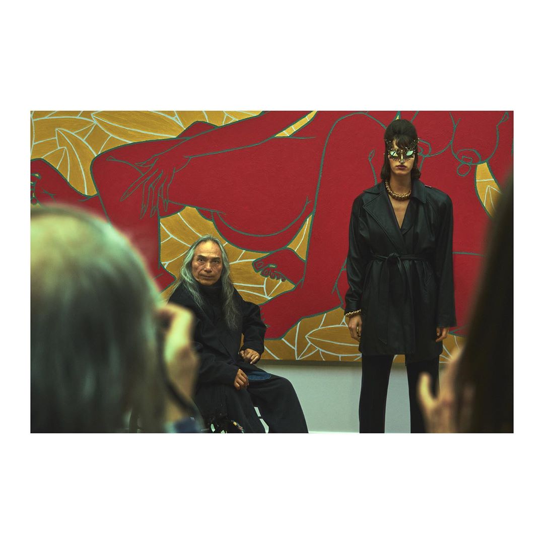 Fashion show featuring a model in a black coat and a seated man in front of an abstract painting with onlookers visible.