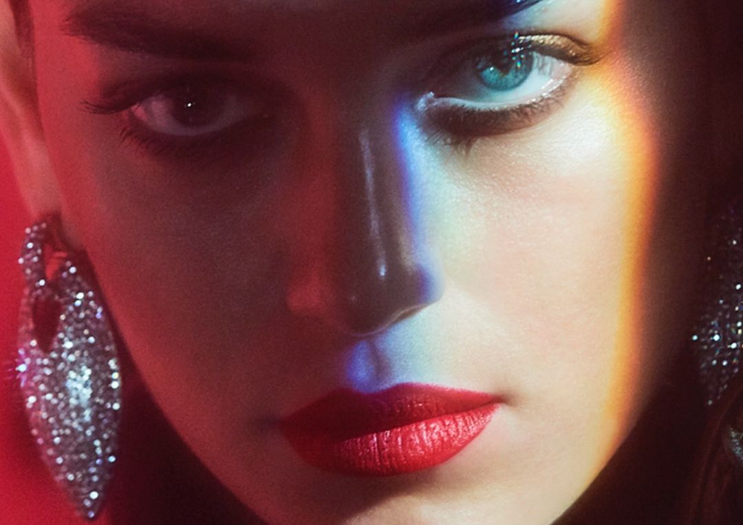 Close-up portrait of a woman with red lipstick and crystal earrings illuminated by a colorful prismatic light stripe across her face.