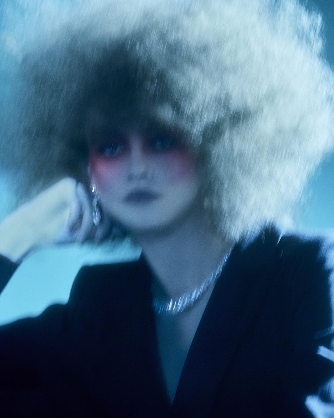 A woman with a large afro hairstyle wears a dark blazer and silver necklace in a blue-toned indoor portrait with soft lighting and blurry focus.