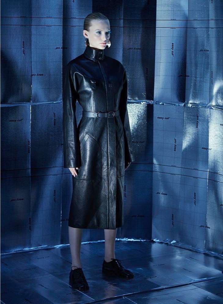 Fashion shot of a woman in a black leather coat and shoes in a blue, tiled photo studio environment. Moody, dramatic lighting.