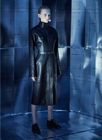 Fashion shot of a woman in a black leather coat and shoes in a blue, tiled photo studio environment. Moody, dramatic lighting.