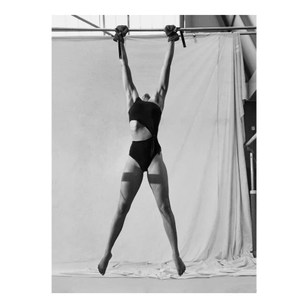 Athletic woman with dark hair hanging from a bar with black wraps in a black bathing suit in a studio with white background.