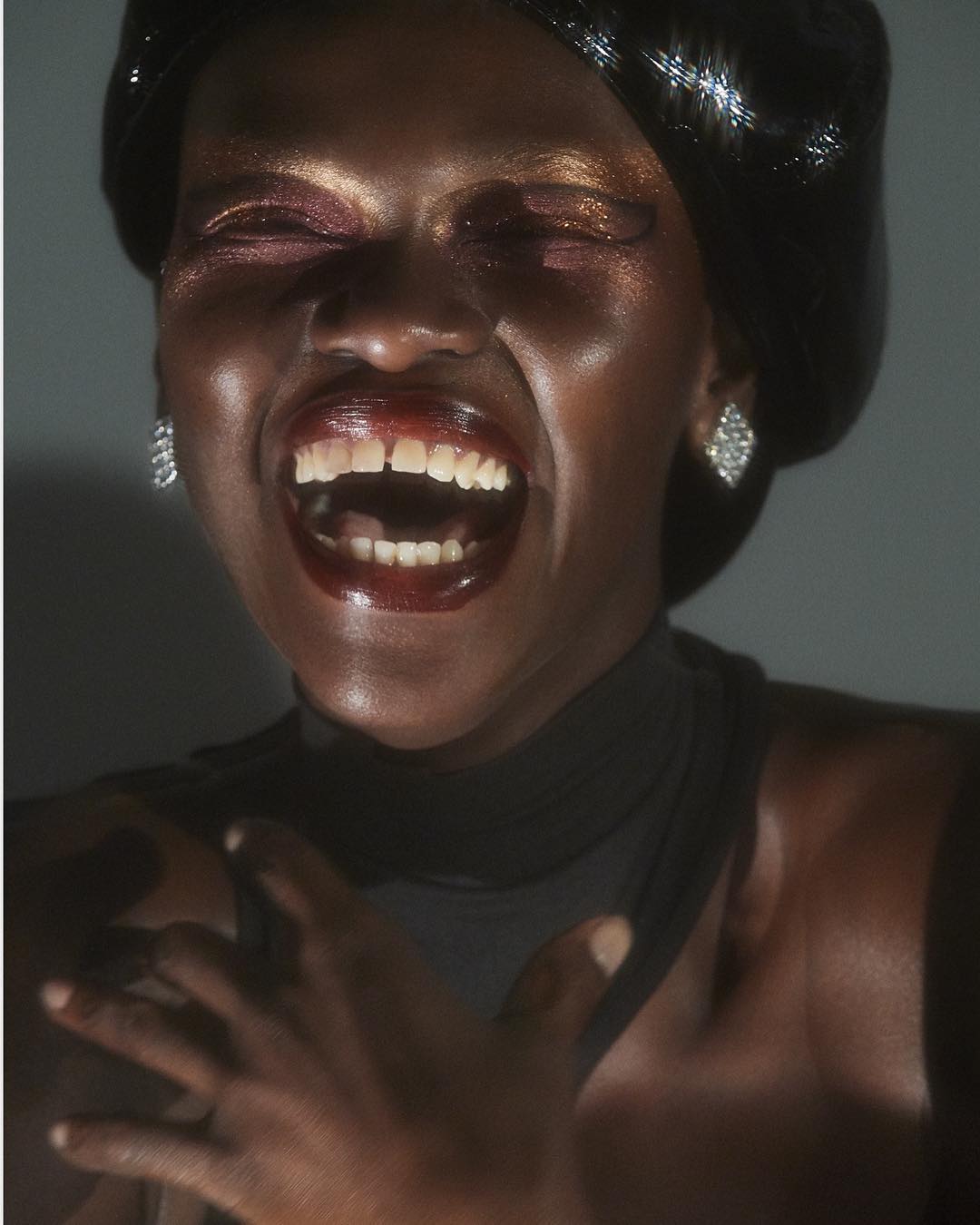 Close-up shot of a stylish Black woman with shimmery gold makeup laughing, wearing a black beret, and diamond earrings against a gray background.