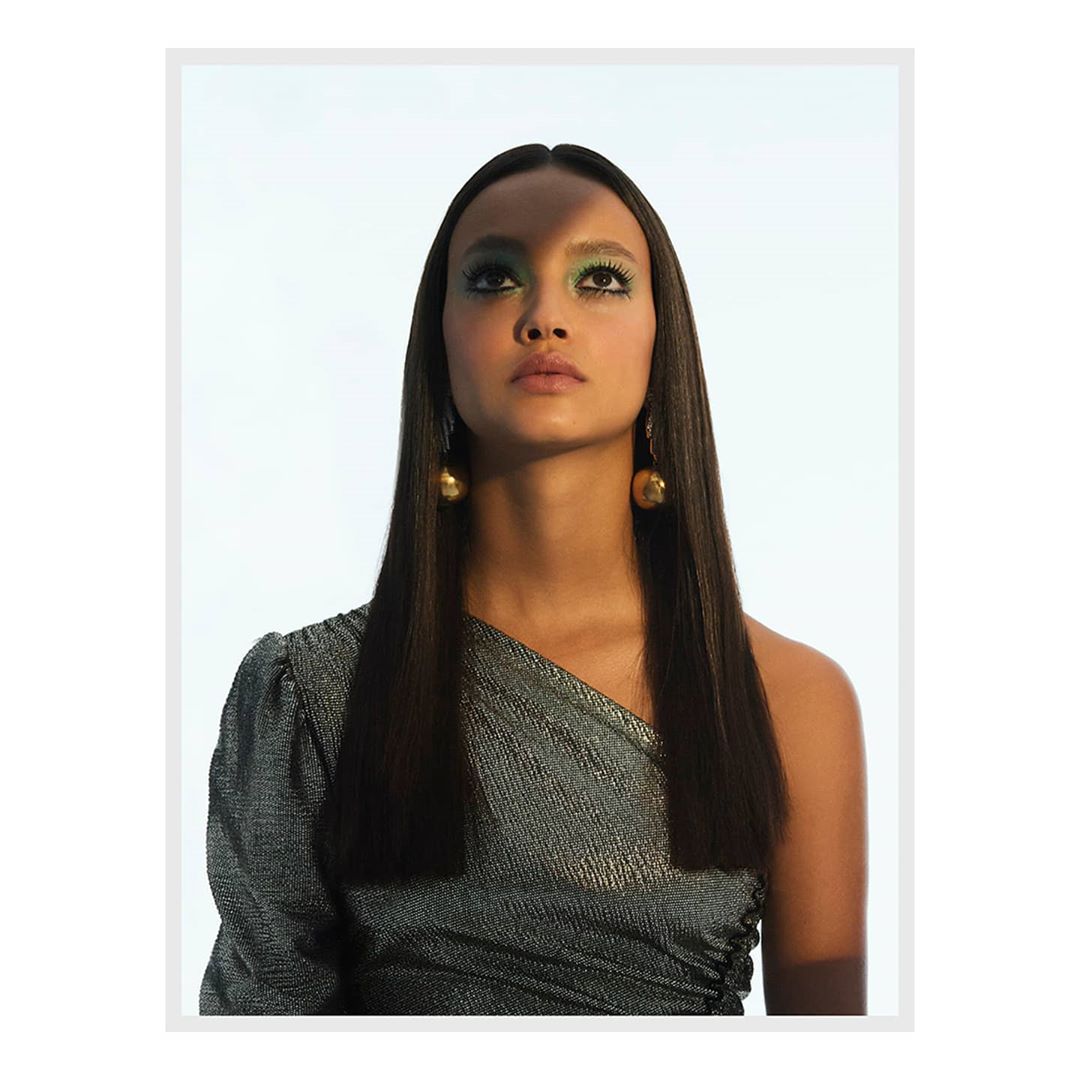 Portrait of a woman with straight dark hair and shimmering green eye makeup looking upward, wearing a one-shoulder top with golden earrings.