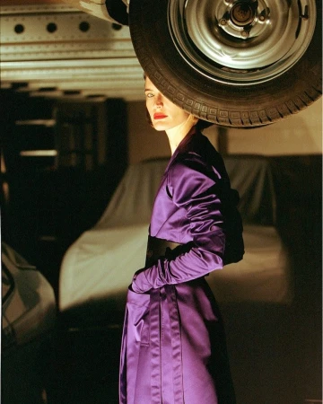 Woman in a glossy purple coat standing beneath a car tire in a dimly lit garage by photographer Lucian Bor