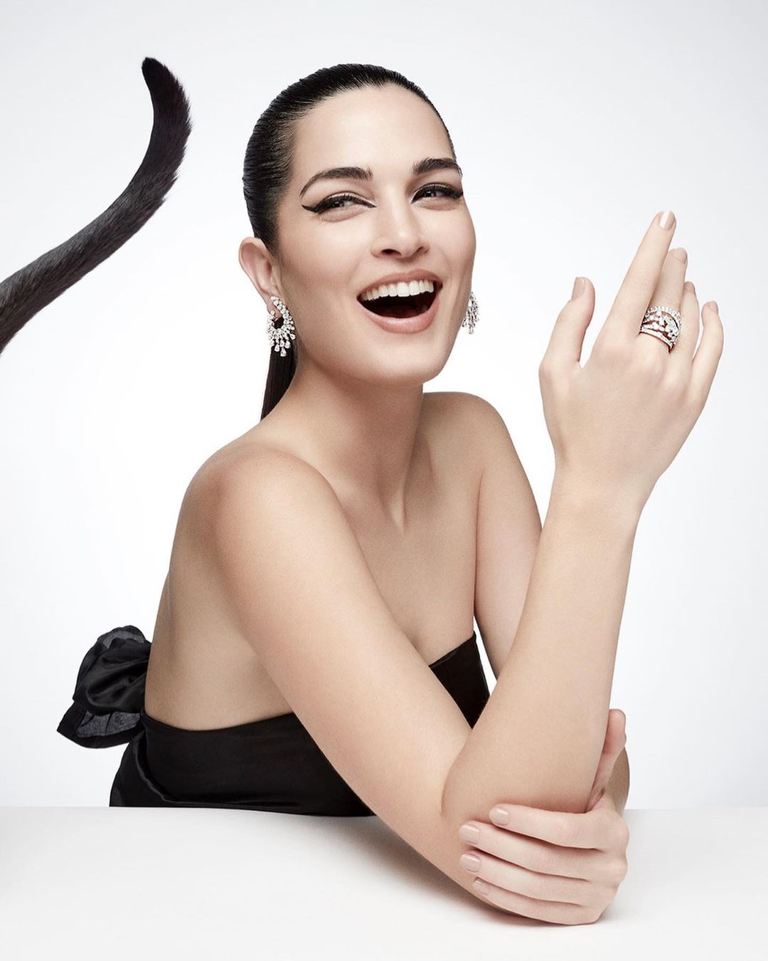 A smiling woman with jewelry poses in a black dress with a decorative bow, set against a bright white background.