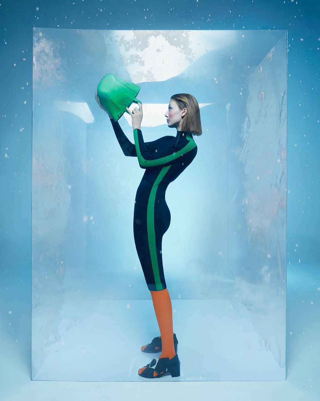 A woman wearing a black and green jumpsuit stands inside a clear cube, holding a green hat, with a light blue and white background.