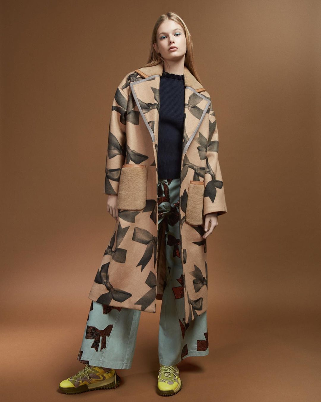 Fashion shoot of a blonde woman in a patterned coat and pants against a tan background, capturing a stylish and editorial vibe.