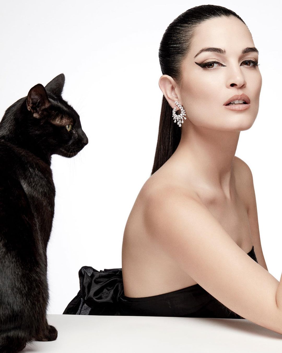 Elegant portrait of an adult female with dark hair and makeup wearing a black dress and large earrings next to a black cat in a studio.