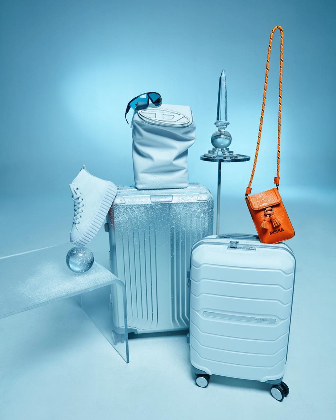 Fashion still life with two suitcases, white sneakers, sunglasses, a handbag, and a small orange purse against a gradient blue background.