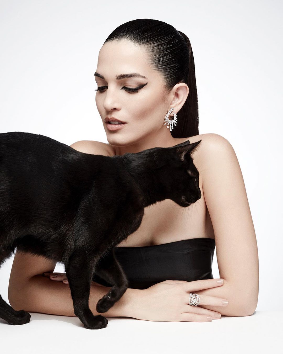 A woman with her hair pulled back poses with a black cat in a studio setting wearing an elegant black dress and jewelry.