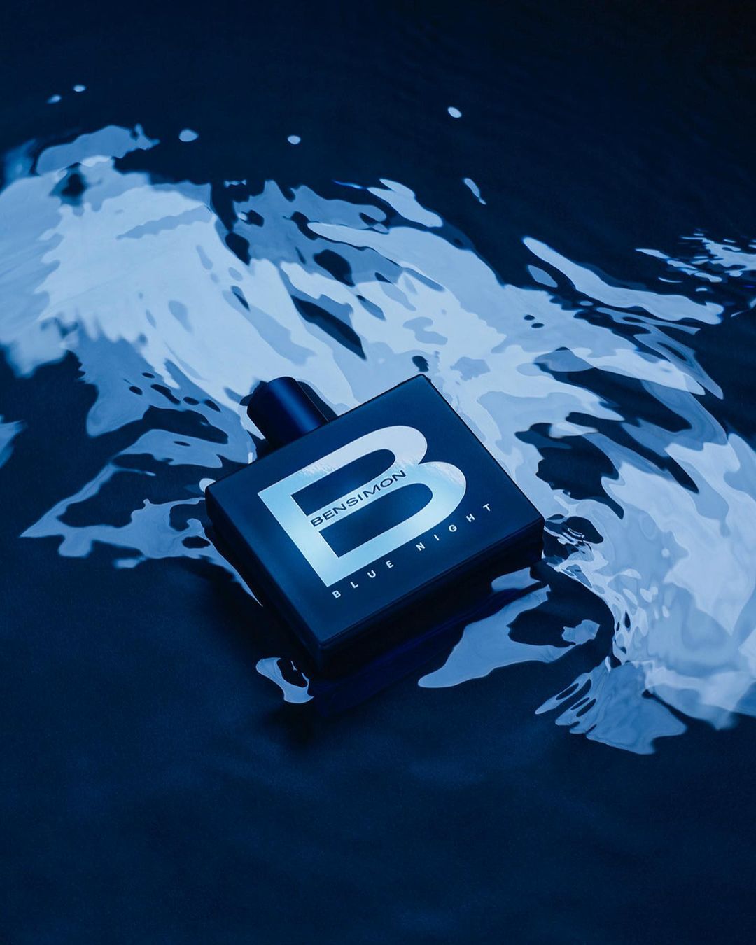 Bensimon Blue Night fragrance bottle is partially submerged in rippled, dark blue water, label facing camera, in a beauty shot.