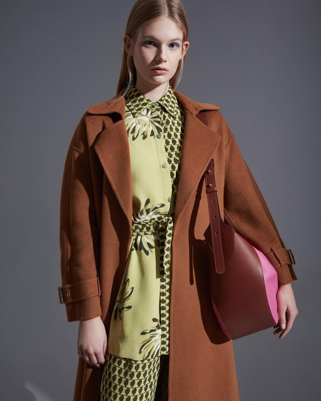A woman in a brown coat, patterned shirt and pants poses in a studio. She carries a two-toned, brown and pink leather shoulder bag.