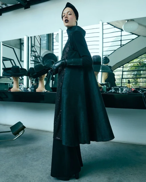 A fashionable woman poses in a dark ensemble featuring a long coat, gloves, and head covering inside a bright, modern room with hats.