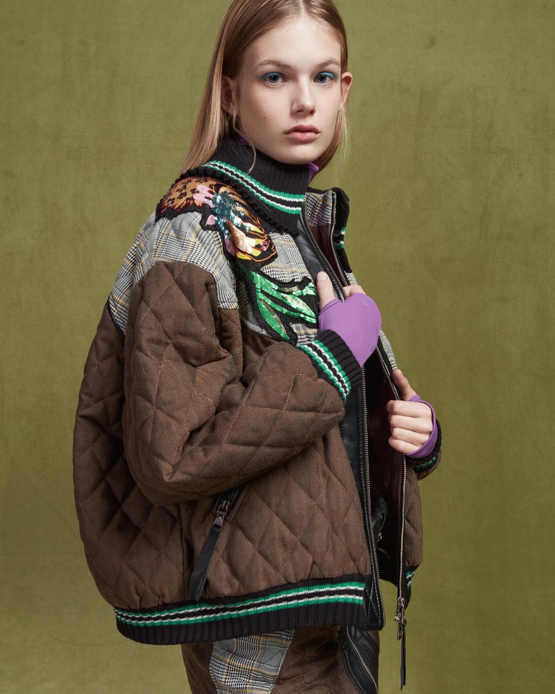 Fashion model wearing a brown quilted jacket with plaid details against a green backdrop in a studio setting with indoor lighting.