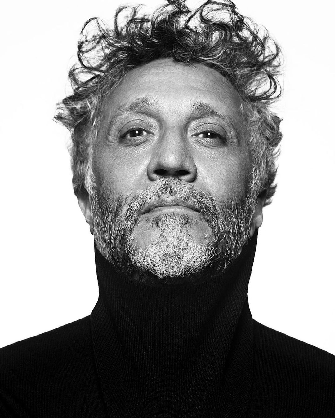 Headshot of an adult male with curly hair and a grey beard, wearing a black turtleneck sweater against a bright white background.