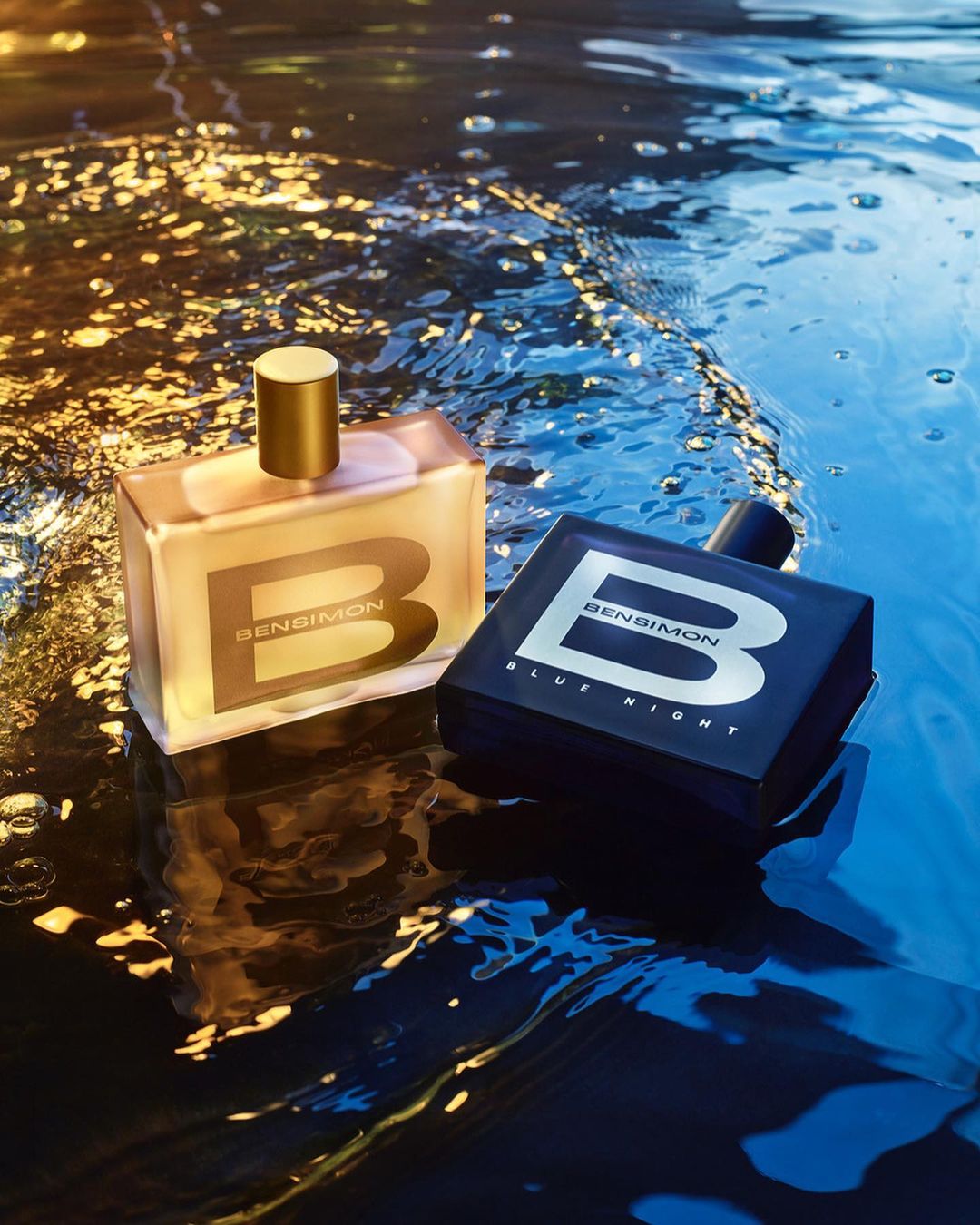 Two Bensimon fragrance bottles, one gold and one blue, are displayed on a reflective wet surface with gold bokeh highlights.