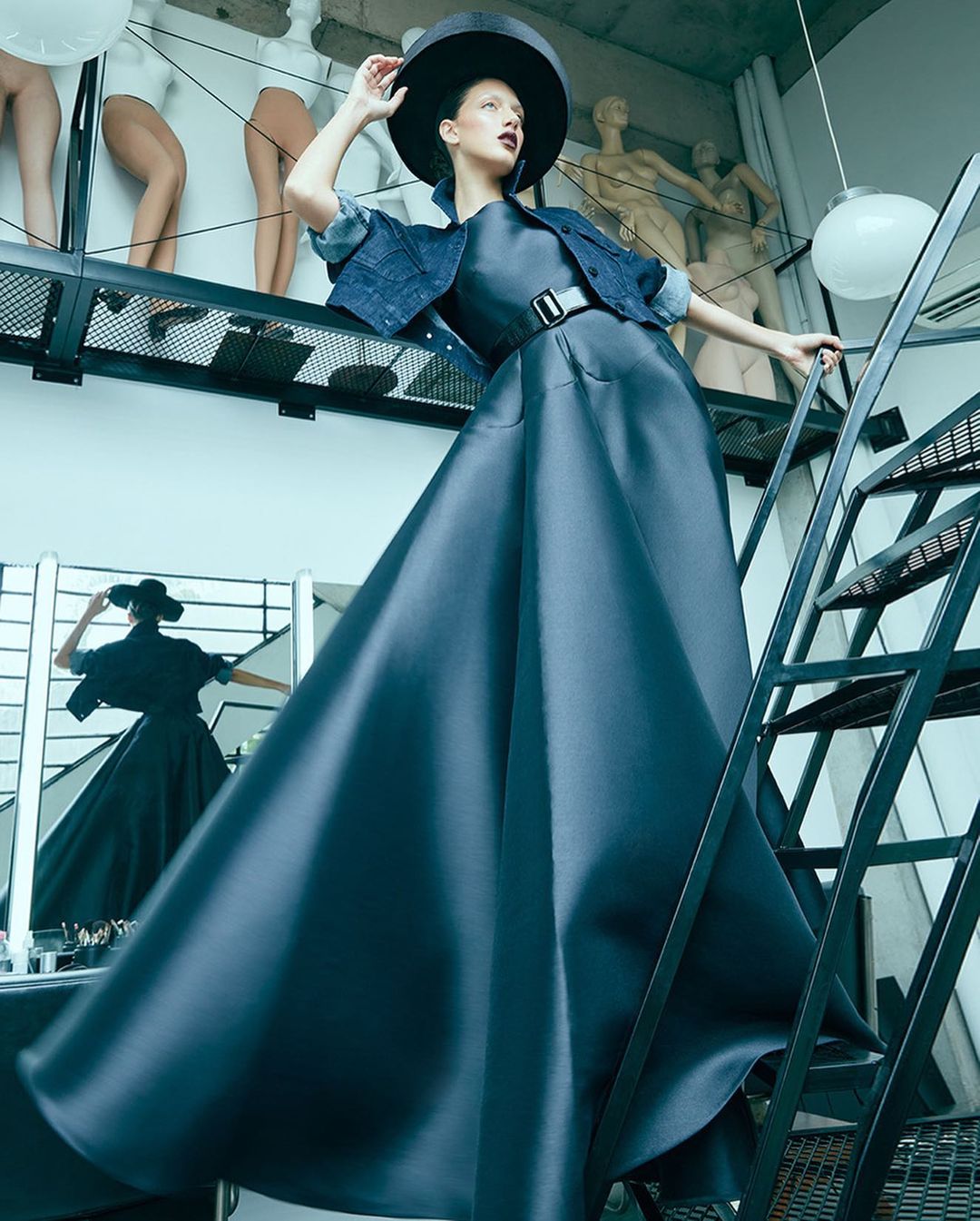 Fashion shot of an adult female model wearing a black hat, dress, and denim jacket on stairs, featuring mannequins in the background.