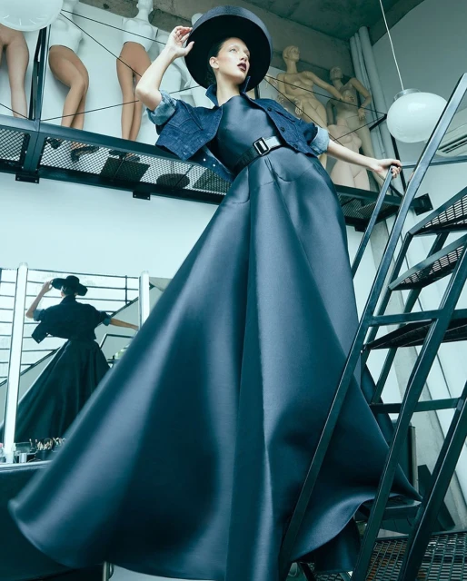 Fashion shot of an adult female model wearing a black hat, dress, and denim jacket on stairs, featuring mannequins in the background.