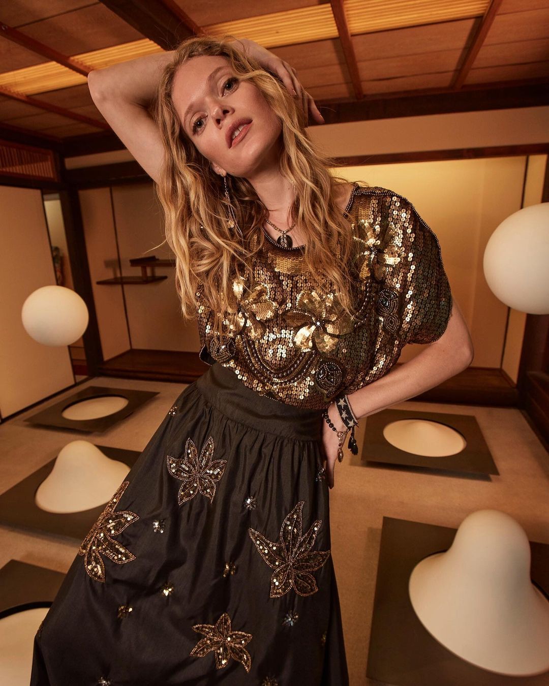 Adult female model wearing a sequined top and embroidered skirt poses in an indoor setting with white globe lighting and dark paneling.