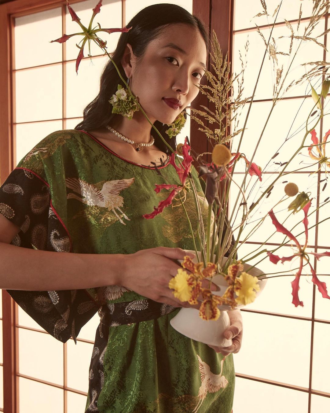 Portrait of an Asian woman in a kimono-style dress holding a floral arrangement, standing in front of a shoji screen with natural light.