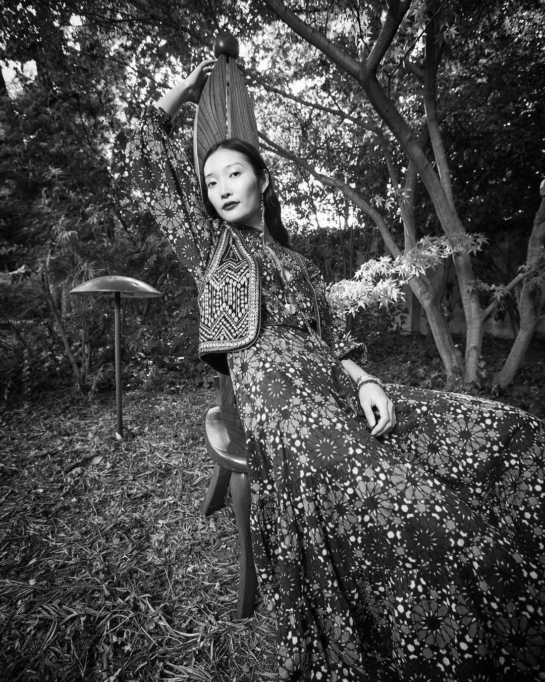 Monochrome portrait of an adult woman in patterned maxi dress and beaded vest, sitting in a chair in a lush garden setting.