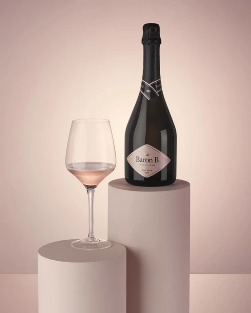 Still life shot of a Baron B rose wine bottle on a pedestal next to a glass of rose wine, against a light pink background.