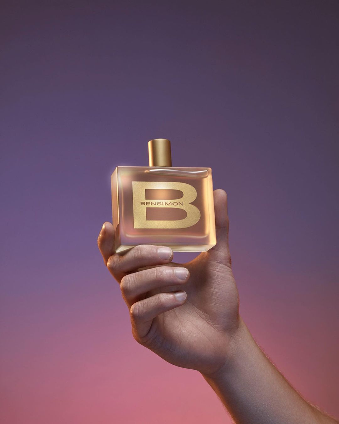 Close-up of a hand holding a Bensimon perfume bottle against a blurred gradient purple and pink background in a beauty shot.
