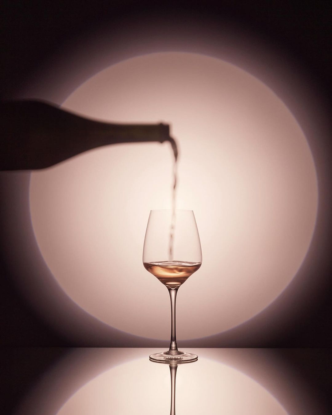 Wine bottle pours liquid into a glass on a reflective surface with soft pink lighting and a circular spotlight in the background.